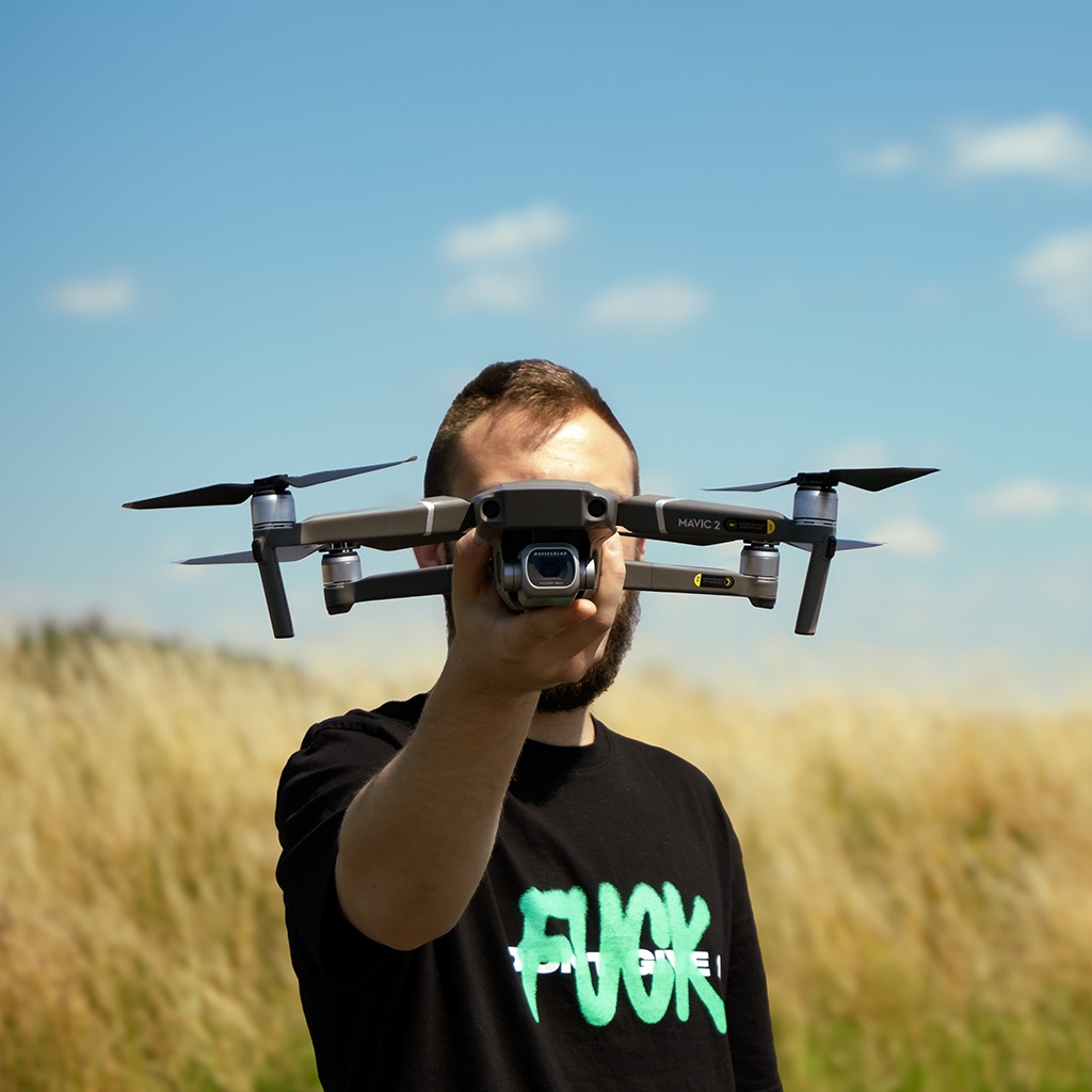 Mężczyzna z brodą trzyma drona Mavic 2 przed twarzą na tle pola i błękitnego nieba. Dron zasłania część twarzy. Widoczna kamera drona. Czarna koszulka z napisem.