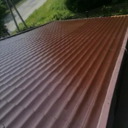 Ciemnobrązowe, metalowe pokrycie dachowe z widocznymi liniami łączeń, sfotografowane z g&oacute;ry, z fragmentem rynny i zielenią w tle.