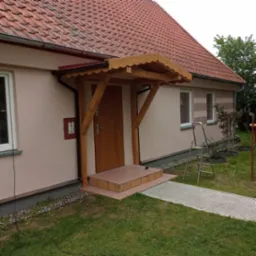 Drewniana, zadaszona weranda przy wejściu do domu z czerwoną dachówką. Widoczne schody wejściowe i fragment trawnika. Jasna elewacja budynku.