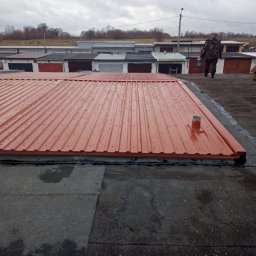 Usługi Ciesielsko- Dekarskie - Sebastian Augustynowicz - Nowe, pomarańczowe krycie dachu garażu z blachy trapezowej, widoczne ślady deszczu. W tle rząd garaży i osoba z pojemnikiem. Widok z góry na płaski dach.