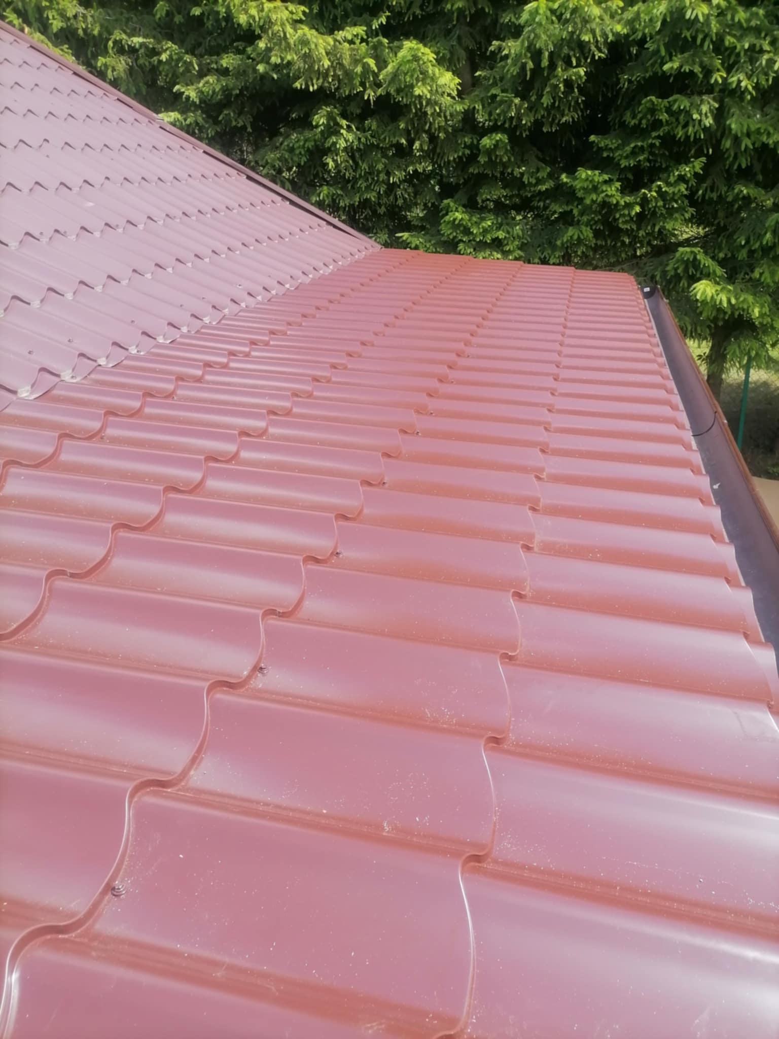 Czerwona blachodachówka na skośnym dachu domu, widok z góry, z zielonymi drzewami w tle. Nowe pokrycie dachowe z wyraźnym wzorem fal.