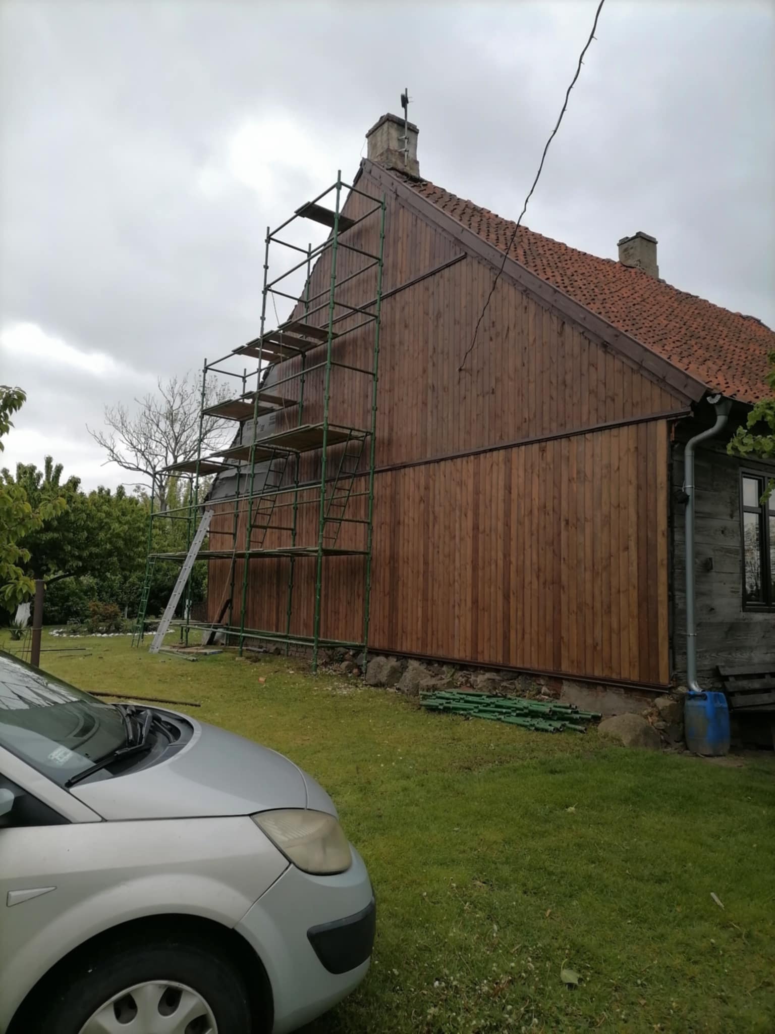 Dom z drewnianą elewacją w trakcie prac konserwacyjnych, widoczne rusztowania i częściowo odnowiona ściana. Czerwona dachówka i fragment srebrnego samochodu.
