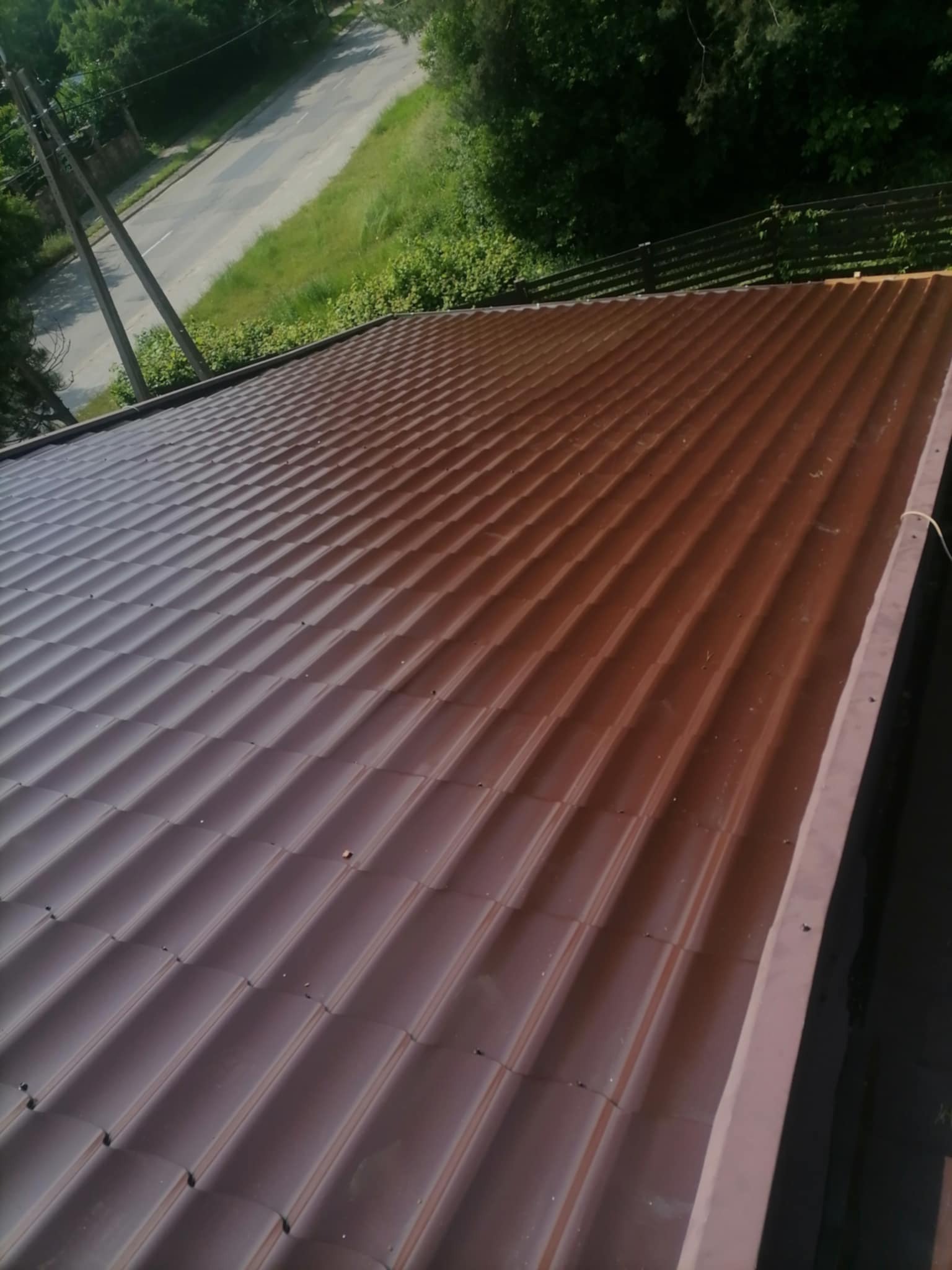 Ciemnobrązowe, metalowe pokrycie dachowe z widocznymi liniami łączeń, sfotografowane z góry, z fragmentem rynny i zielenią w tle.