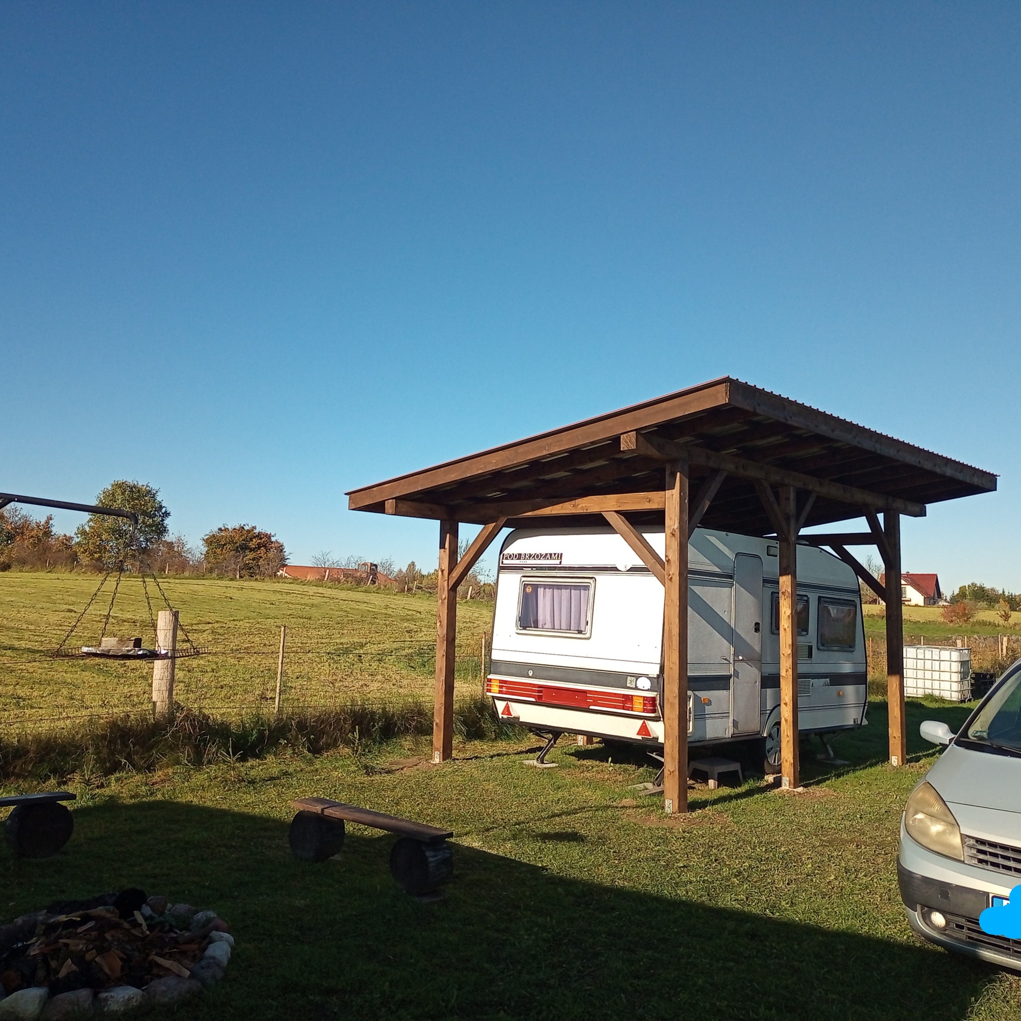 Kempingowy zadaszony wóz na zielonej działce, widok z boku. Drewniana konstrukcja zadaszenia, w tle drzewa i błękitne niebo. Częściowo widoczny samochód.