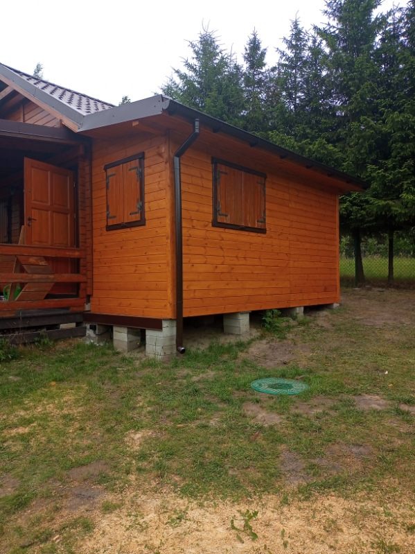 Drewniany domek letniskowy z brązowymi okiennicami i dachem, otoczony zielenią. Widoczne rynny i podmurówka z betonowych bloczków. Ujęcie z poziomu gruntu.