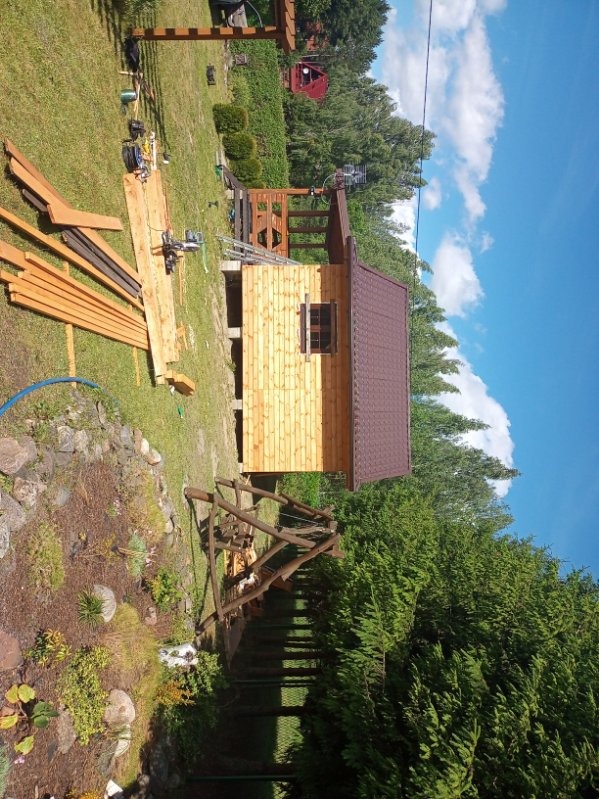 Budowa drewnianej altany z brązowym dachem w ogrodzie, widoczne narzędzia i materiały budowlane, częściowo zmontowana pergola w tle, słoneczny dzień.