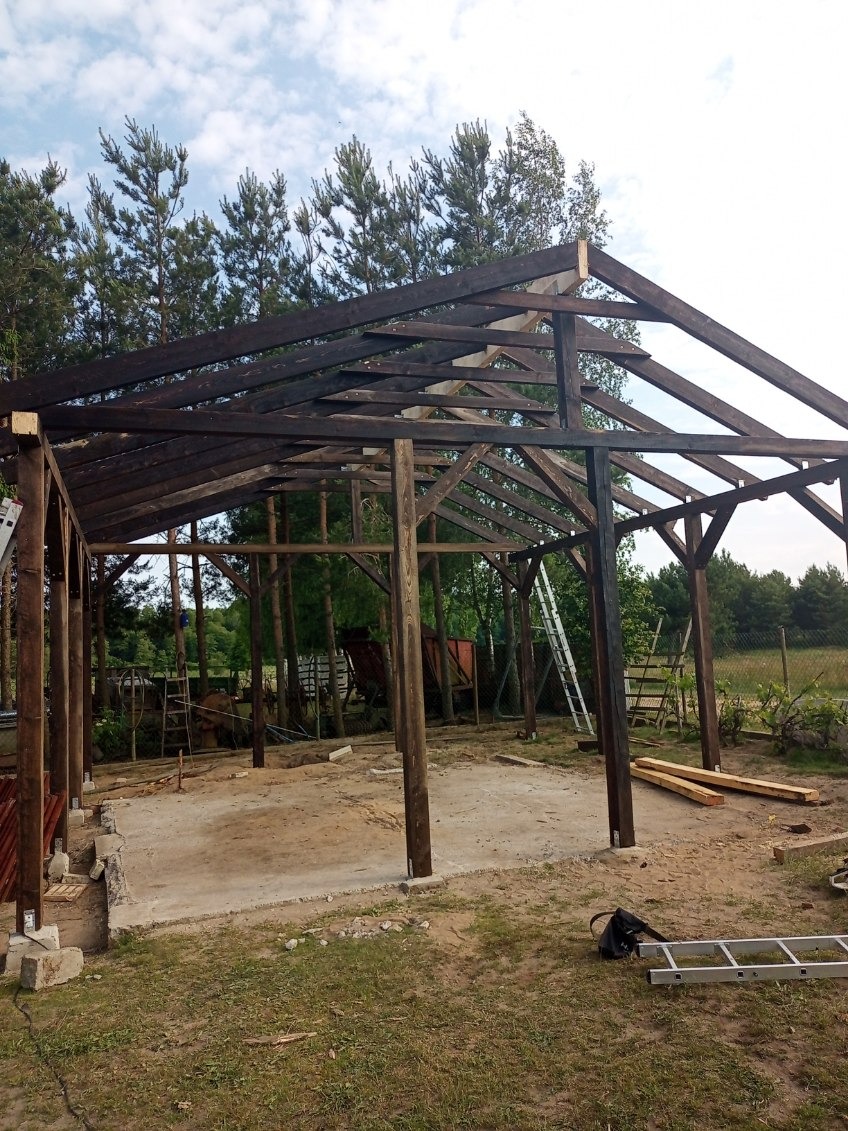 Konstrukcja drewniana z ciemnego drewna w trakcie budowy, widoczne słupy i więźba dachowa, w tle zieleń drzew i niebo. Etap budowy z widocznymi narzędziami.