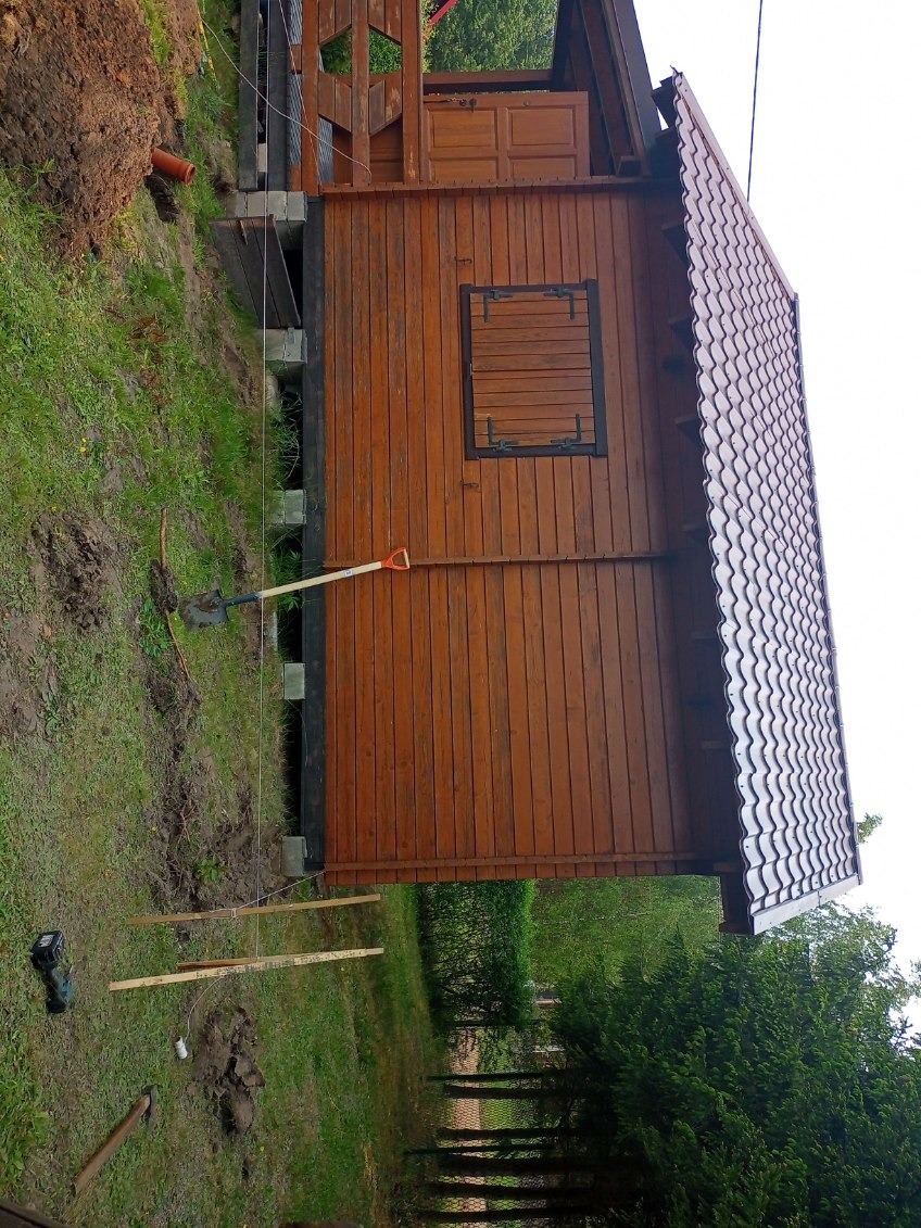 Drewniana altana z brązowym dachem z blachodachówki, widok z boku. Na trawie leżą narzędzia i deski, co sugeruje prace wykończeniowe.