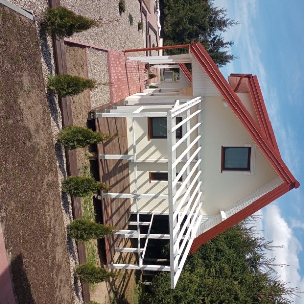 Biała altana z drewnianym tarasem i czerwonym dachem, otoczona zielenią i kamieniami, widziana z boku pod kątem, w słoneczny dzień.