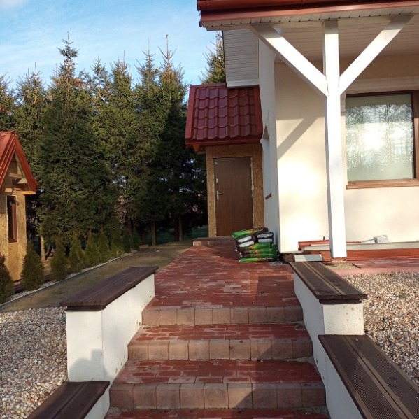 Ceglane schody wejściowe z siedziskami prowadzą do domu z brązowym dachem i drewnianymi drzwiami. W tle iglaki i budynek gospodarczy.