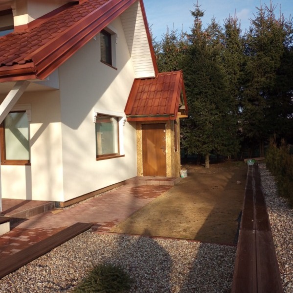 Biały dom z brązowym dachem i wejściem z drewnianą obudową. Ułożona kostka brukowa i rabaty z kamykami. W tle wysokie, zielone drzewa iglaste.