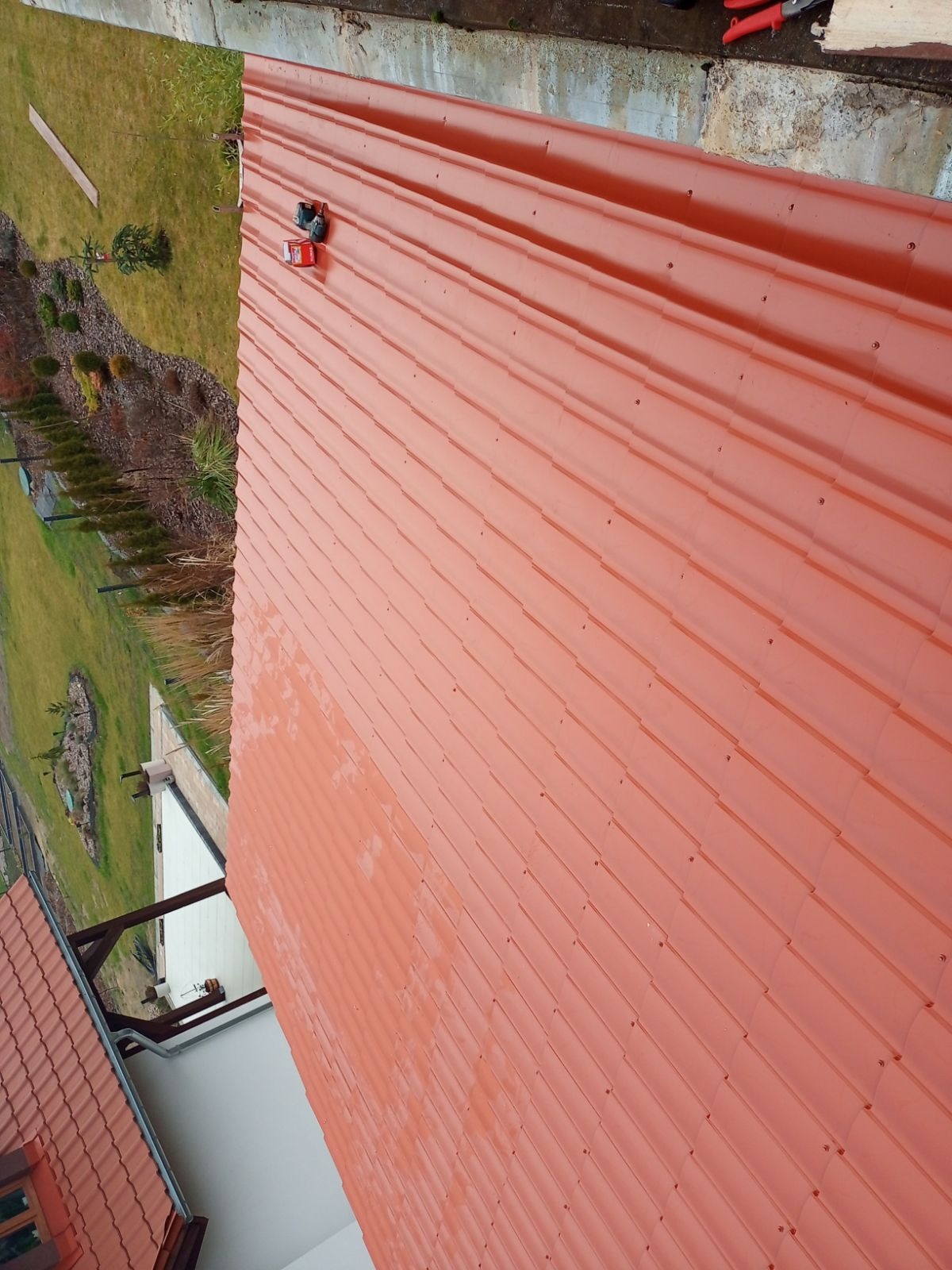 Czerwona blachodachówka na skośnym dachu domu, widok z góry, narzędzia dekarskie leżą na powierzchni. W tle zieleń ogrodu i fragment sąsiedniego dachu.