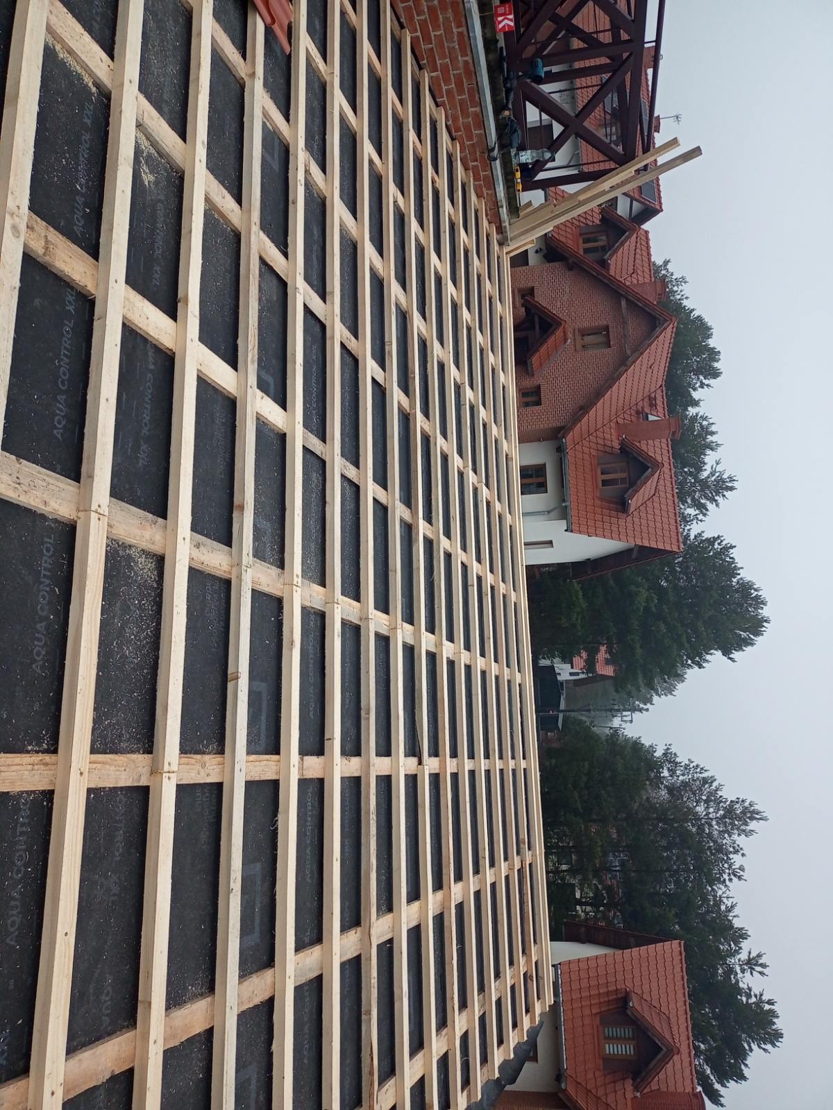 Konstrukcja drewniana na czarnej membranie dachowej, widok z góry na tle budynków z czerwonymi dachami. Etap budowy dachu.