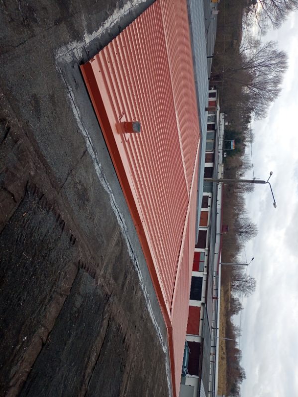 Czerwony, blaszany dach garażu z widocznymi obróbkami blacharskimi i uszczelnieniami, na tle innych garaży i drzew. Ujęcie z góry, lekko z boku.