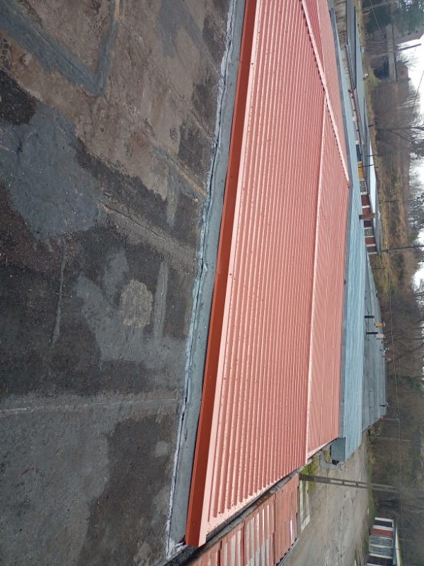 Czerwony dach z blachy trapezowej na budynku przemysłowym, widok z góry, z widocznymi detalami łączeń i obróbek blacharskich. Część dachu pokryta papą.