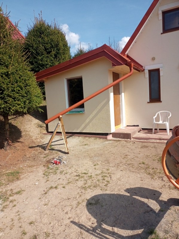 Montaż rynny w Lidzbarku Warmińskim. Rynna w kolorze ceglastym, przymocowana do budynku z jasną elewacją. Widoczne drzewa iglaste i fragment betoniarki.