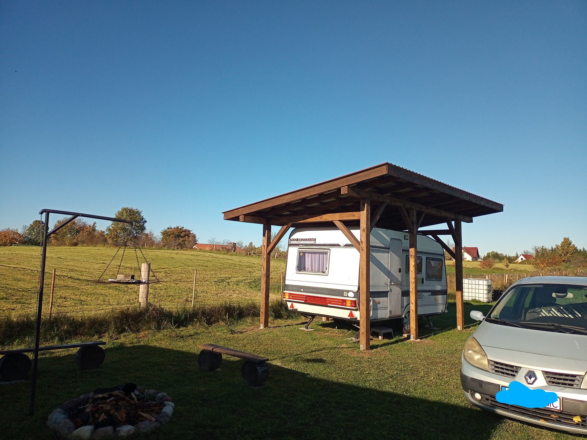 Pergola z drewnianą konstrukcją chroniąca przyczepę kempingową na zielonej działce. W tle łąka i drzewa, z boku zaparkowany samochód.