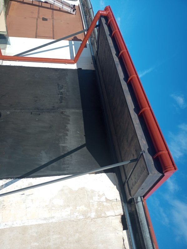 Czerwona rynna dachowa zamontowana na krawędzi dachu z papy, widok z dołu na błękitne niebo. Elementy mocujące rynnę widoczne na tle ściany budynku.