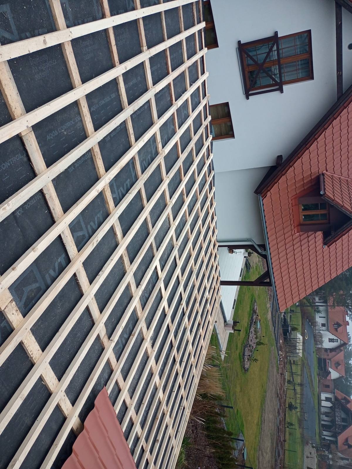 Konstrukcja dachu z łatami i membraną w trakcie budowy, widok z góry na dom z czerwoną dachówką i zielony ogród w tle. Elementy drewniane w jasnym odcieniu.