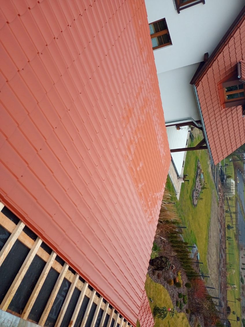 Czerwona blachodachówka na nowym dachu domu jednorodzinnego, widok z góry na konstrukcję więźby. W tle ogród i fragment drugiego dachu.