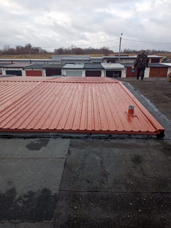 Nowe, pomarańczowe krycie dachu garażu z blachy trapezowej, widoczne ślady deszczu. W tle rząd garaży i osoba z pojemnikiem. Widok z góry na płaski dach.