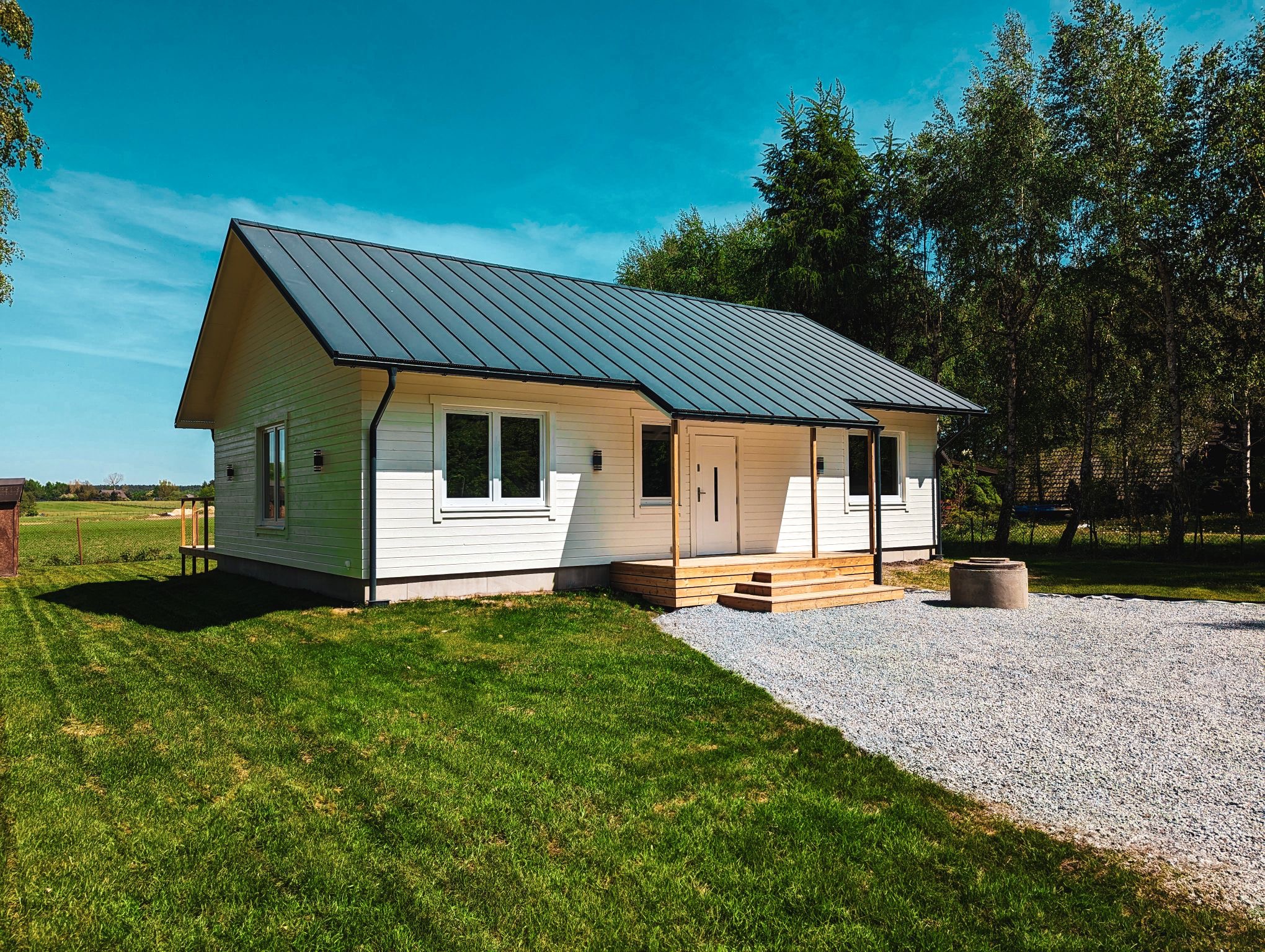 Gotowy dom szkieletowy z ciemnym dachem w Sierakówek. Biała elewacja, drewniane schody wejściowe, otoczenie: trawnik i drzewa. Ujęcie w słoneczny dzień.