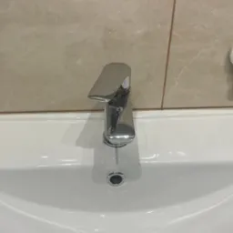 Biała umywalka Keramin z chromowaną baterią w łazience z beżowymi płytkami. Umywalka czysta, po sprzątaniu, z widocznym odpływem i uchwytem na ścianie.