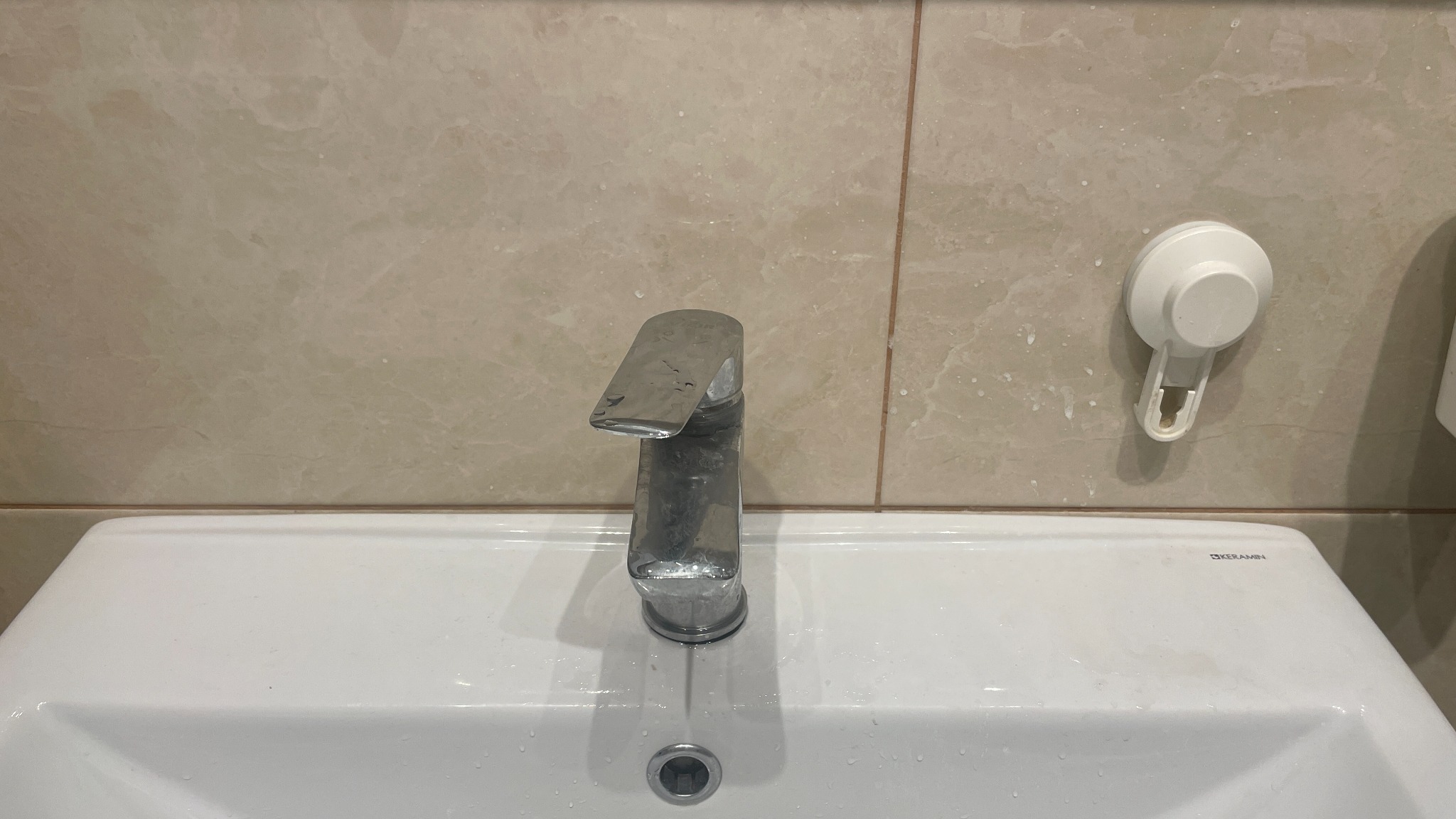 Biała umywalka z chromowaną baterią i śladami zaschniętej wody na beżowych płytkach, obok uchwyt na szczotkę. Widoczne zabrudzenia po remoncie.