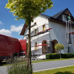 Dom w trakcie ocieplania, widoczne rusztowania, pracownik przy oknie na poddaszu, czerwony samochód dostawczy i drzewo na pierwszym planie.