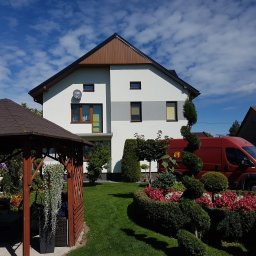 USŁUGI BUDOWLANE SŁAWOMIR KALISIAK - Dwukondygnacyjny dom z białą elewacją i szarym pasem, brązowym dachem, otoczony zielonym trawnikiem i ozdobnymi krzewami, z czerwonym vanem w tle.