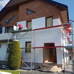 USŁUGI BUDOWLANE SŁAWOMIR KALISIAK - Dwóch pracowników na rusztowaniu montuje białe płyty izolacyjne na elewacji domu z ceglanym piętrem. Widoczne narzędzia i materiały budowlane.