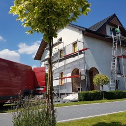 USŁUGI BUDOWLANE SŁAWOMIR KALISIAK - Dom w trakcie prac elewacyjnych: rusztowania, pracownik przy oknie, częściowo otynkowana ściana, czerwony bus w tle, błękitne niebo z chmurami.