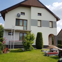 USŁUGI BUDOWLANE SŁAWOMIR KALISIAK - Dwukondygnacyjny dom po częściowym malowaniu elewacji w Grabicy. Widoczne rusztowanie, drabina i narzędzia malarskie na trawniku. Świeża, jasna kolorystyka.