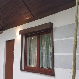 USŁUGI BUDOWLANE SŁAWOMIR KALISIAK - Elewacja domu z oknem z brązowymi roletami i dekoracyjnym malowaniem w poziome pasy szarości. Widoczny fragment dachu i drzwi wejściowe.