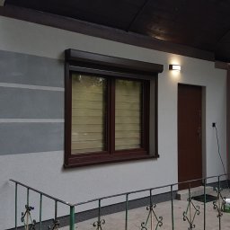 USŁUGI BUDOWLANE SŁAWOMIR KALISIAK - Dom po malowaniu elewacji z szarymi pasami, brązowe okna z roletami, oświetlenie zewnętrzne. Widoczne barierki i fragment ogrodu.