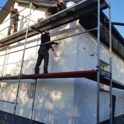 USŁUGI BUDOWLANE SŁAWOMIR KALISIAK - Tynkowanie elewacji domu jednorodzinnego z rusztowania. Pracownicy w trakcie nakładania tynku. Widoczne narzędzia i materiały budowlane. Częściowo otynkowana ściana.