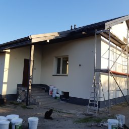 USŁUGI BUDOWLANE SŁAWOMIR KALISIAK - Dom jednorodzinny po malowaniu elewacji na biało, z widocznymi rusztowaniami i pracownikiem na nich. W tle błękitne niebo i farby w wiaderkach.
