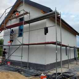 USŁUGI BUDOWLANE SŁAWOMIR KALISIAK - Trzech pracowników na rusztowaniu tynkuje elewację domu jednorodzinnego na biało. Okno zabezpieczone folią. Widoczne wiaderka z zaprawą i narzędzia.