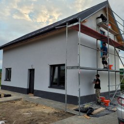 USŁUGI BUDOWLANE SŁAWOMIR KALISIAK - Trzech pracowników na rusztowaniu tynkuje elewację domu jednorodzinnego na szaro. Widoczne wiaderka z zaprawą i narzędzia. Dom z czarną podmurówką i stolarką okienną.