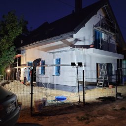 USŁUGI BUDOWLANE SŁAWOMIR KALISIAK - Dom jednorodzinny w trakcie tynkowania elewacji zewnętrznej. Na rusztowaniach pracownicy. Wieczorne oświetlenie budowy. Widoczne narzędzia i materiały budowlane.