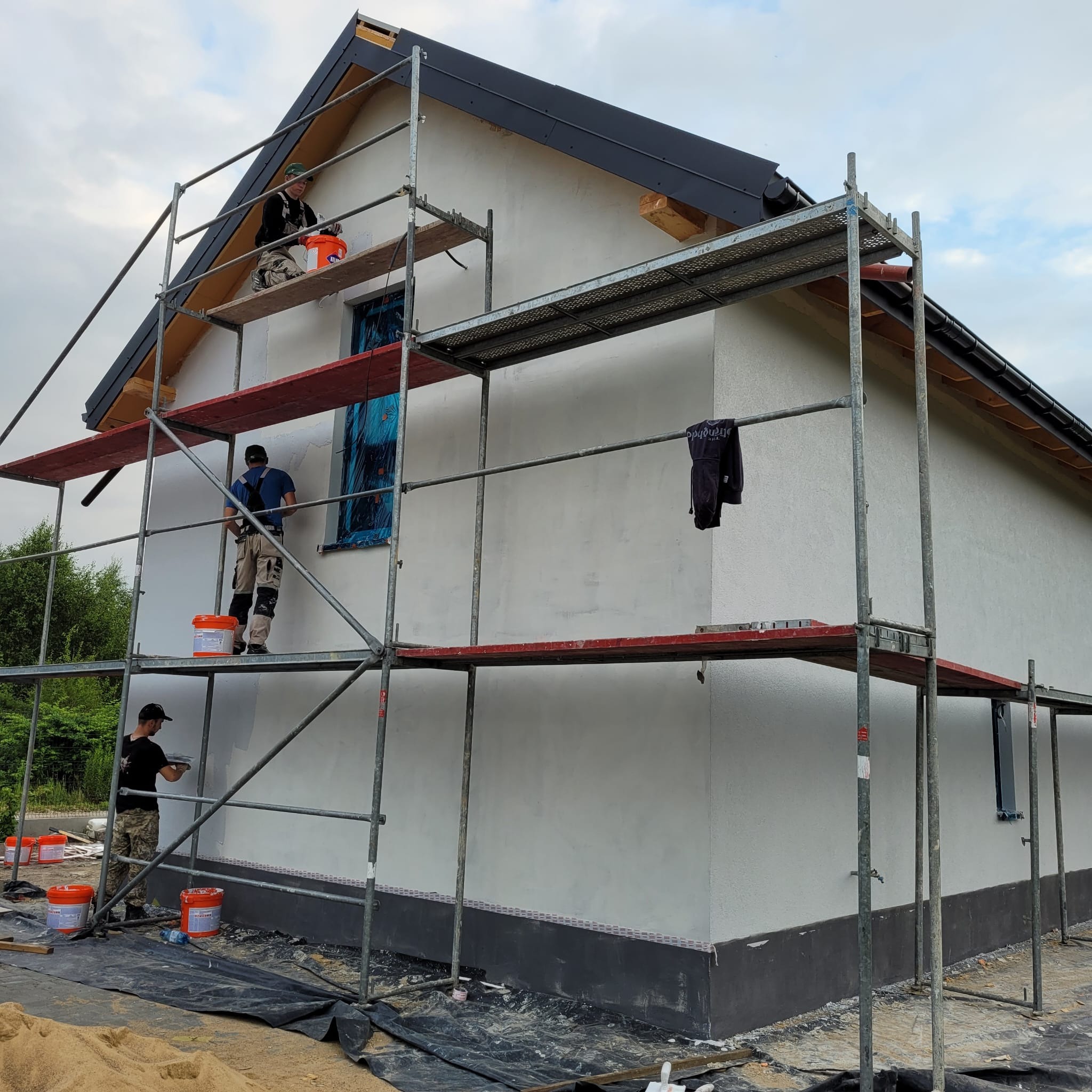 Praca wre: Trzech mężczyzn na rusztowaniu tynkuje elewację domu jednorodzinnego. Widoczne wiaderka z zaprawą, narzędzia i zabezpieczone okno. Elewacja w jasnym odcieniu.