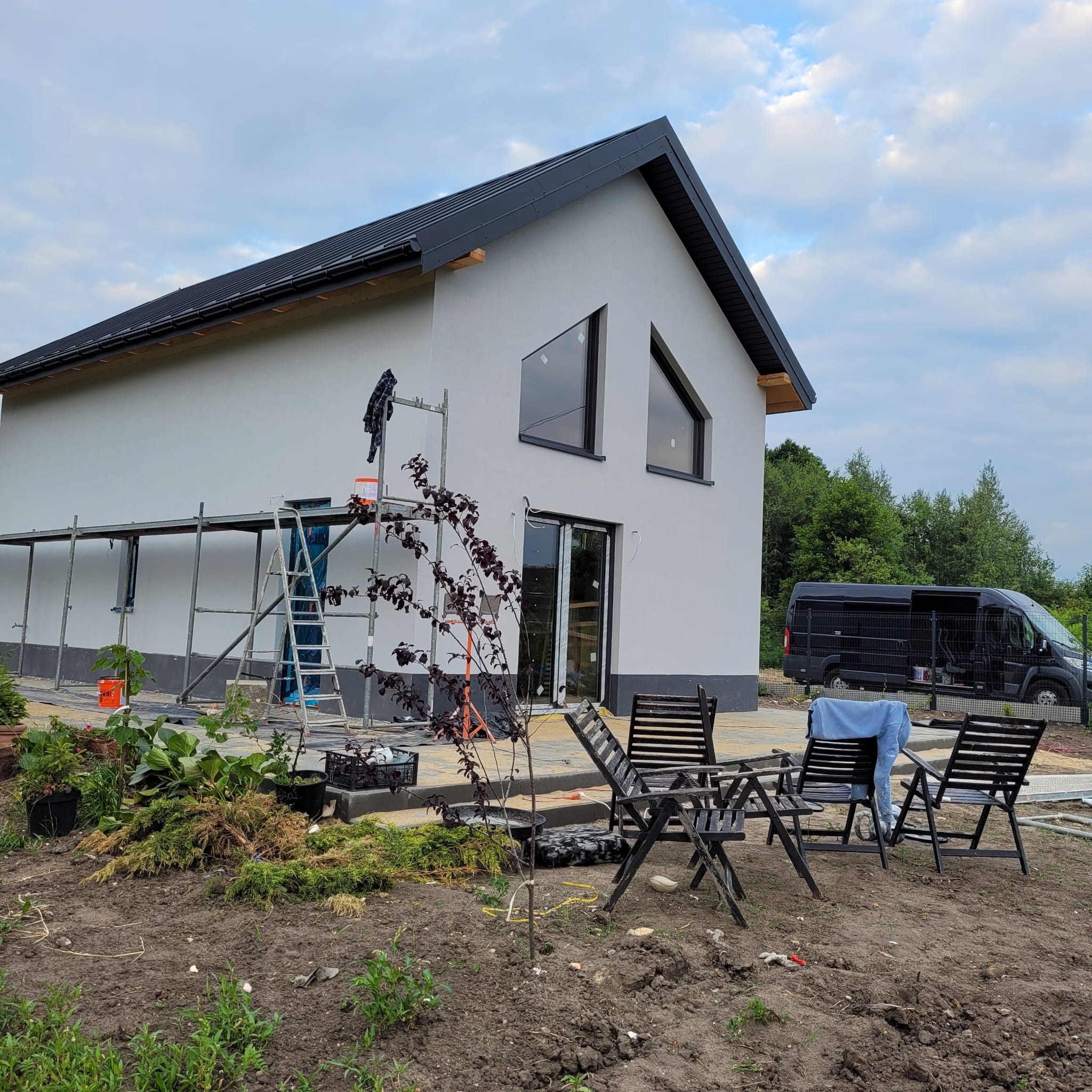 Nowoczesny dom z szarą elewacją po tynkowaniu, z czarną stolarką okienną i drabiną na rusztowaniu. W tle czarny van i zieleń. Widoczne meble ogrodowe.