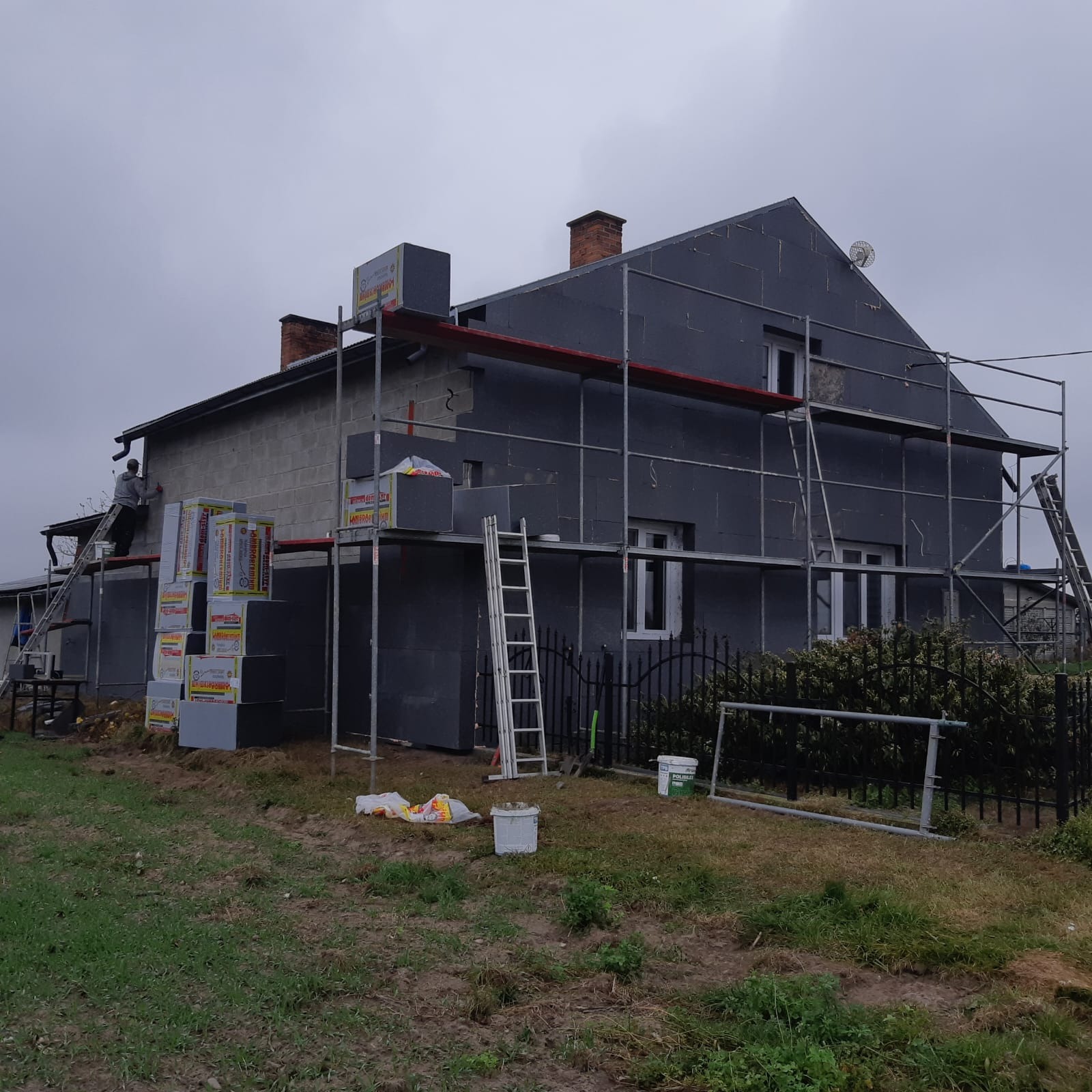 Dom w trakcie ocieplania styropianem grafitowym, widoczne rusztowania i pracownik na drabinie. Część elewacji już ocieplona, reszta w trakcie.