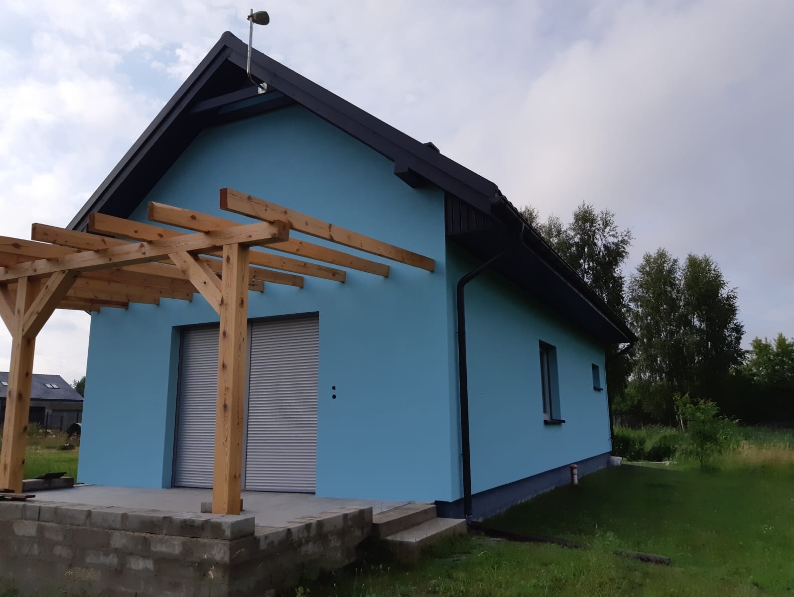 Błękitny dom z czarnym dachem i drewnianą pergolą nad garażem. Świeżo pomalowana elewacja w letnim krajobrazie. Widoczne rolety garażowe i betonowe schody.