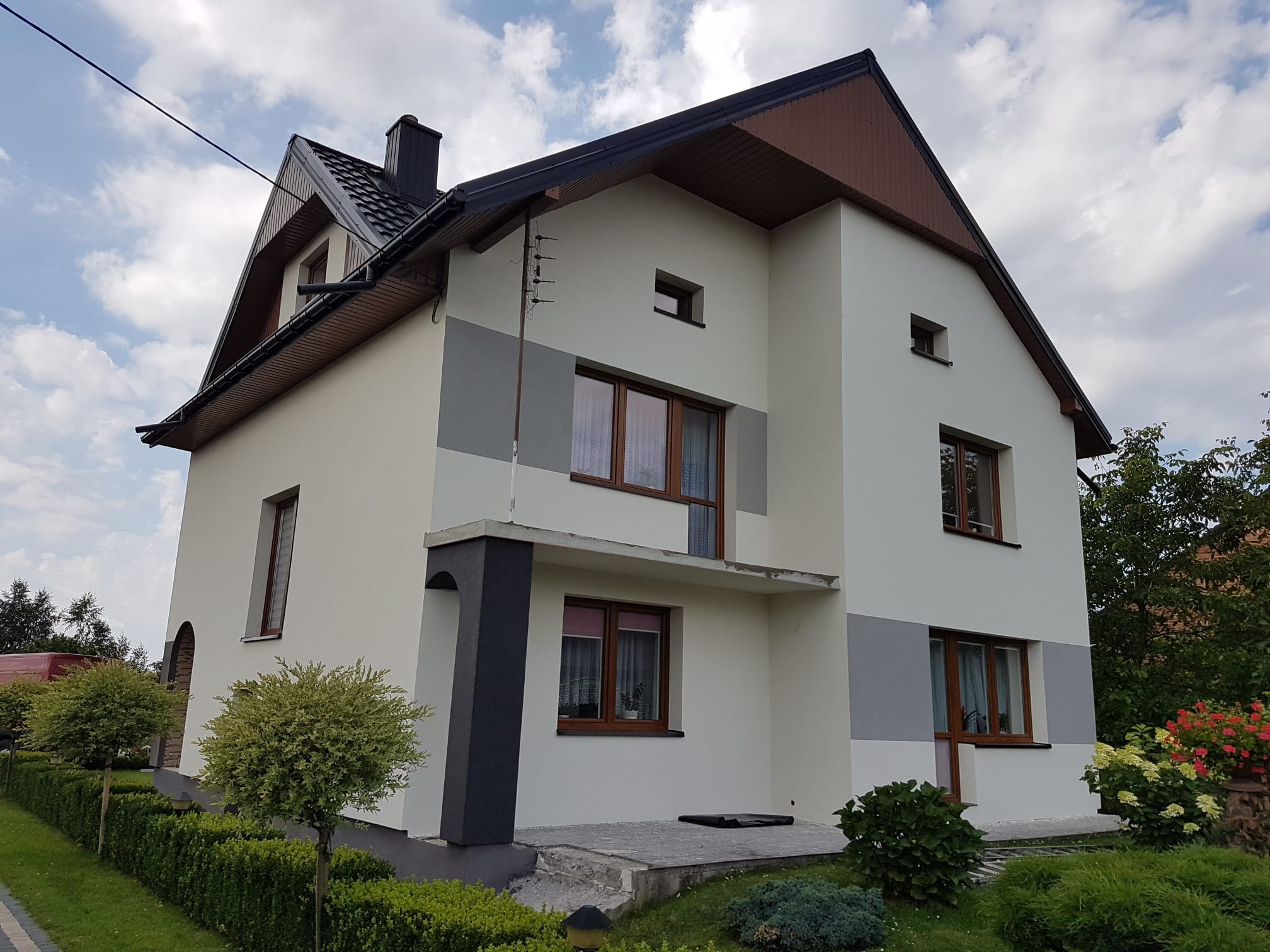 Dwukondygnacyjny dom z kremową elewacją, szarymi akcentami i brązowym dachem, otoczony zielenią. Widoczna antena na ścianie.