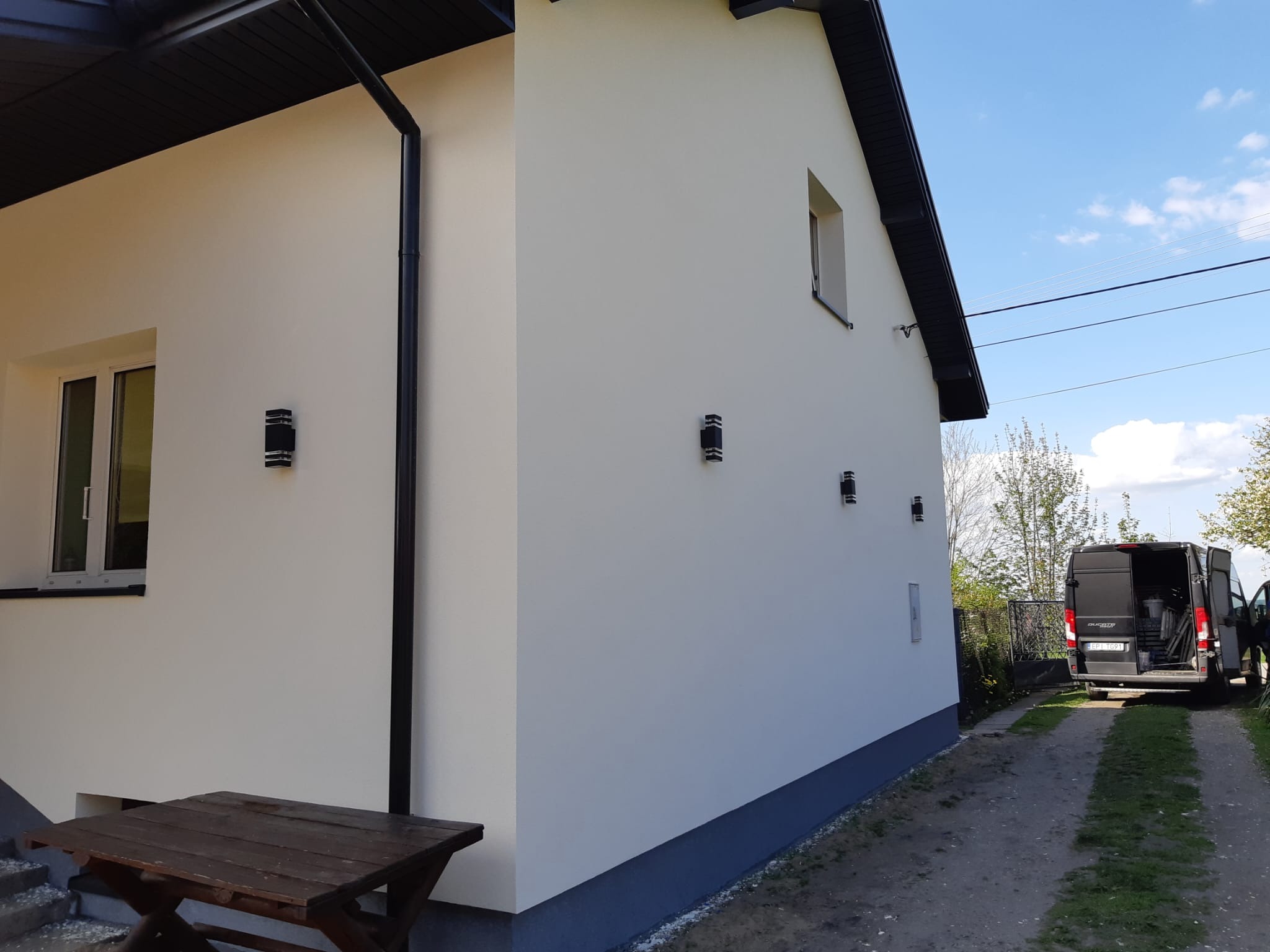 Jasna elewacja domu z ciemnymi akcentami (rynny, dach), oświetlona promieniami słońca. Widoczny fragment stołu i zaparkowany samochód dostawczy.