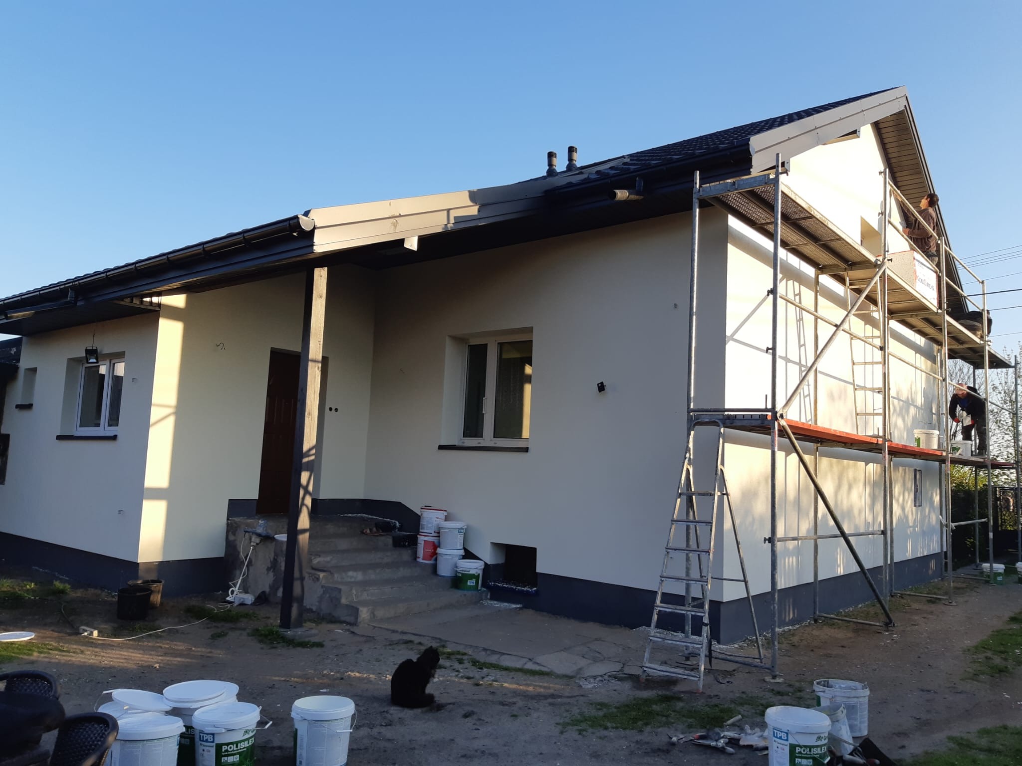 Dom po malowaniu elewacji na biało, widoczne rusztowania i puszki z farbą.  Czarny kot siedzi na ziemi przed domem.