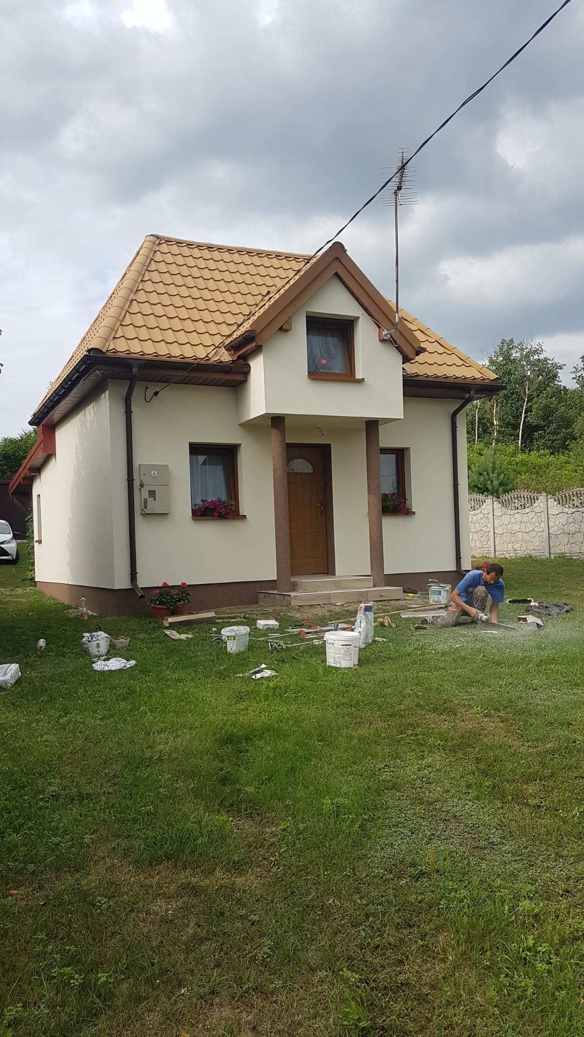 Dom jednorodzinny po tynkowaniu elewacji, z widocznymi detalami wykończenia i pracownikiem przygotowującym teren. Widoczne puszki i narzędzia.