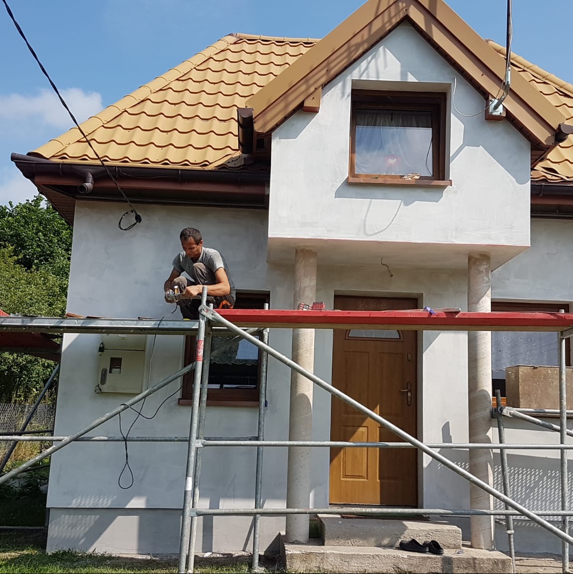 Pracownik na rusztowaniu obrabia elewację domu z beżowym dachem. Widoczne przygotowania do tynkowania lub malowania. Dom z kolumnami przy wejściu.