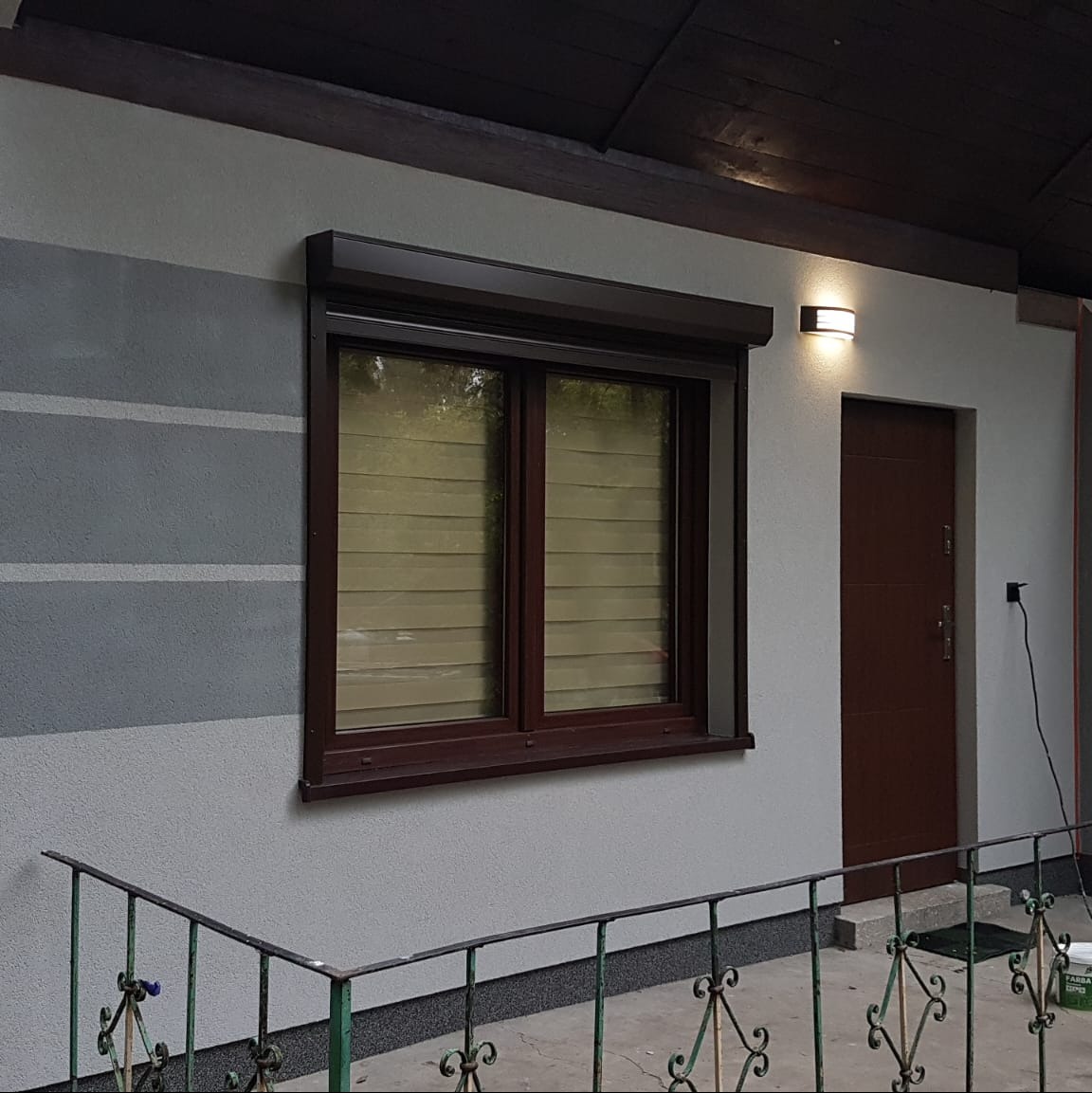 Elewacja domu z oknem z brązową ramą i roletą. Ściana pomalowana w poziome pasy szarości. Obok brązowe drzwi wejściowe i oświetlenie zewnętrzne.